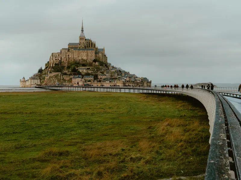 Le Mont-Saint-Michel sous un ciel gris ; la passerelle courbe y mène des promeneurs, le long d’un pré.