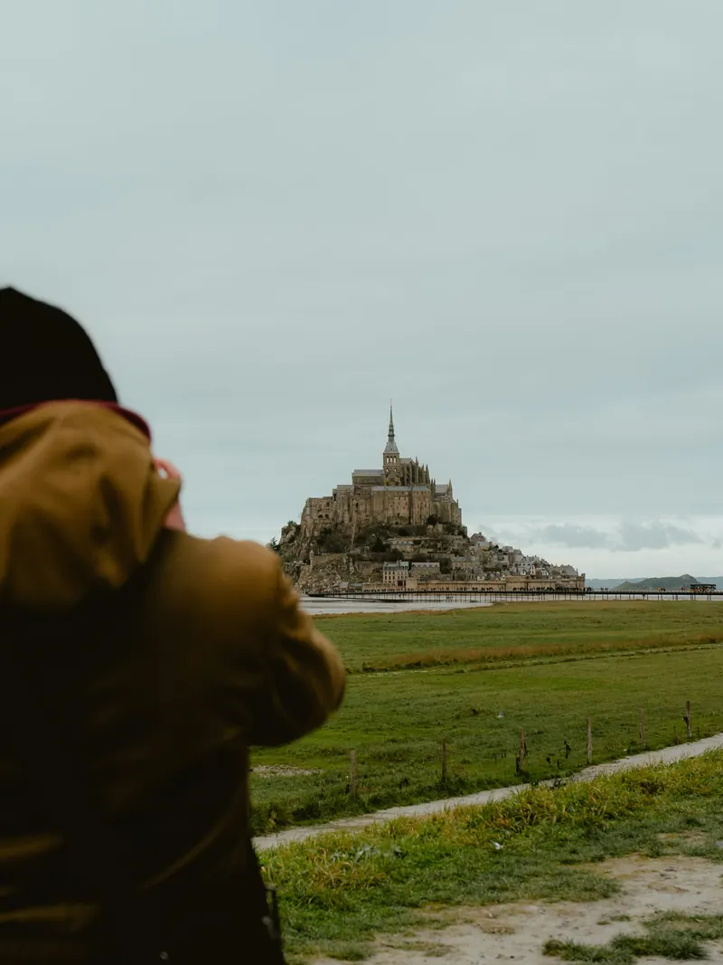 Au premier plan, de dos et flou, une personne en veste ocre et bonnet sombre, les bras levés face au Mont-Saint-Michel, au loin au-delà des prés.