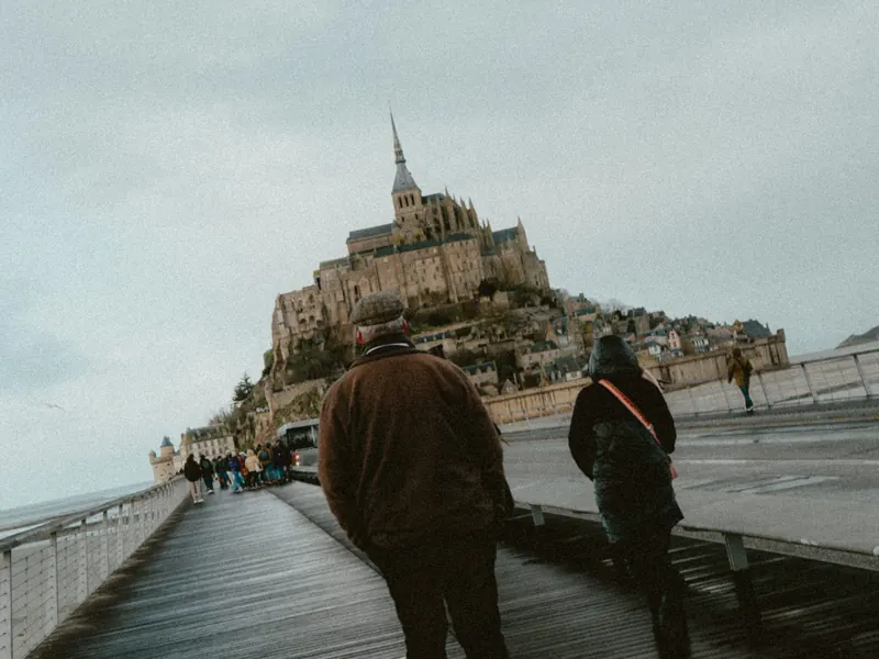 Deux promeneurs en manteau, vus de dos, avancent sur la passerelle mouillée vers le Mont-Saint-Michel.