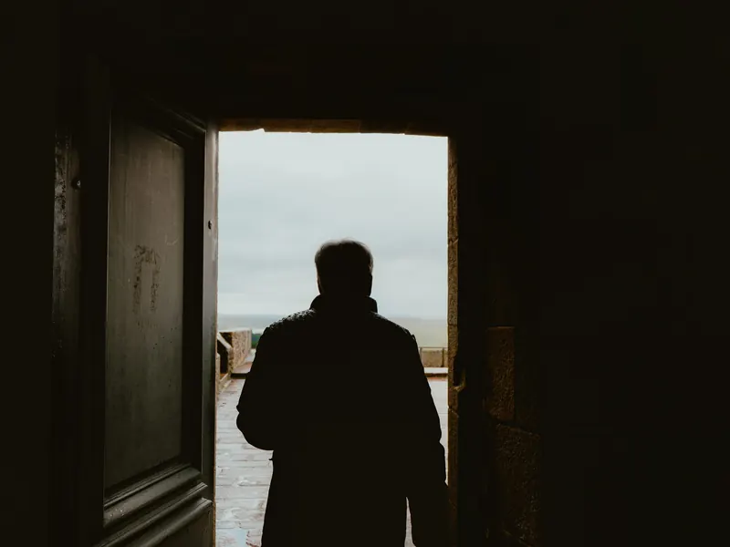Un homme en silhouette, vu de dos, franchit une porte sombre qui donne sur une terrasse de pierre et un ciel blanc.