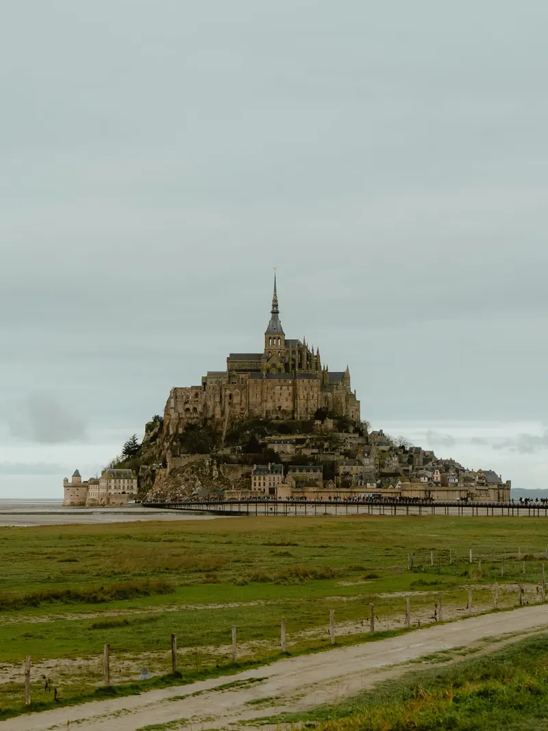 Le Mont-Saint-Michel sous un ciel gris, vu depuis les prés ; au premier plan, un chemin de terre longé d’une clôture.