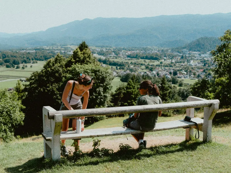 Deux personnes à un banc de bois blanc, sur une hauteur herbeuse : l’une se penche, l’autre est assise face à une vallée habitée et à des collines.