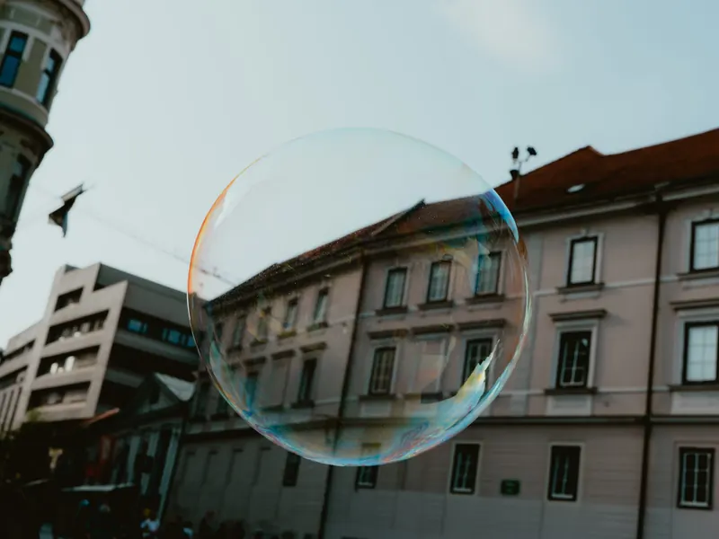 Une grande bulle de savon irisée flotte dans une rue, devant la façade claire d’un immeuble ancien et un bâtiment moderne.
