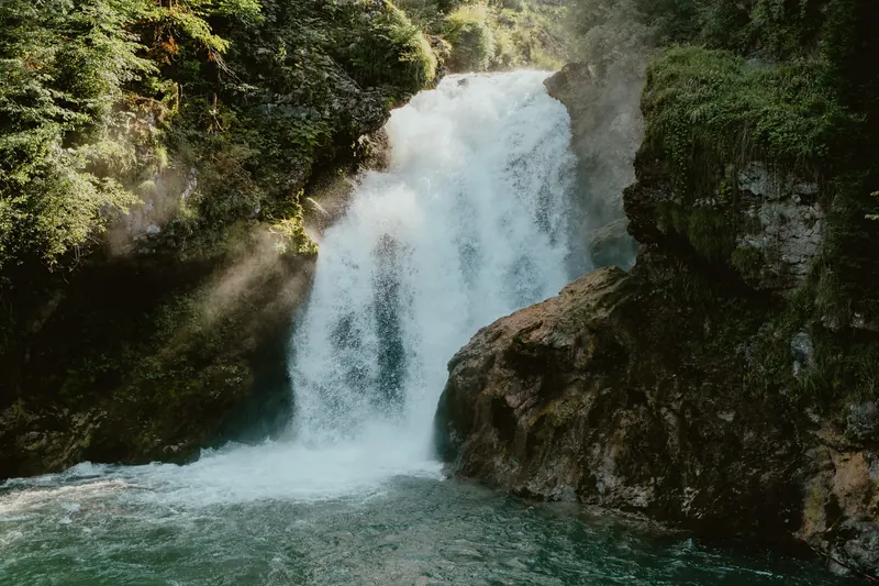 Une cascade tombe entre des rochers couverts de mousse dans un bassin, traversée par un rayon de soleil.
