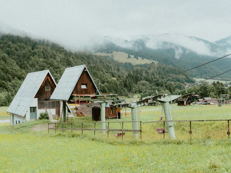 Deux chalets de bois au toit pointu et les pylônes d’une remontée mécanique dans un pré, devant des montagnes prises dans la brume.