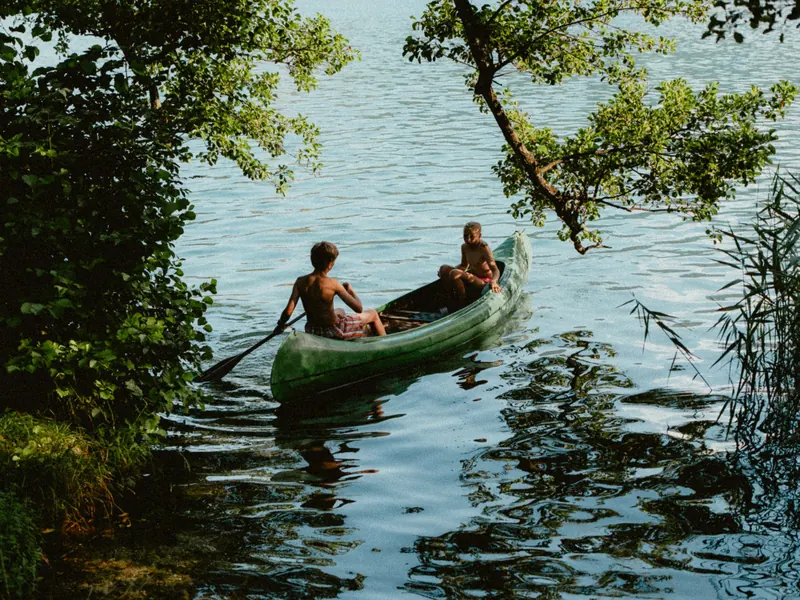 Deux enfants dans un canoë vert pagaient sur un lac, sous les branches d’un arbre penché au-dessus de l’eau.