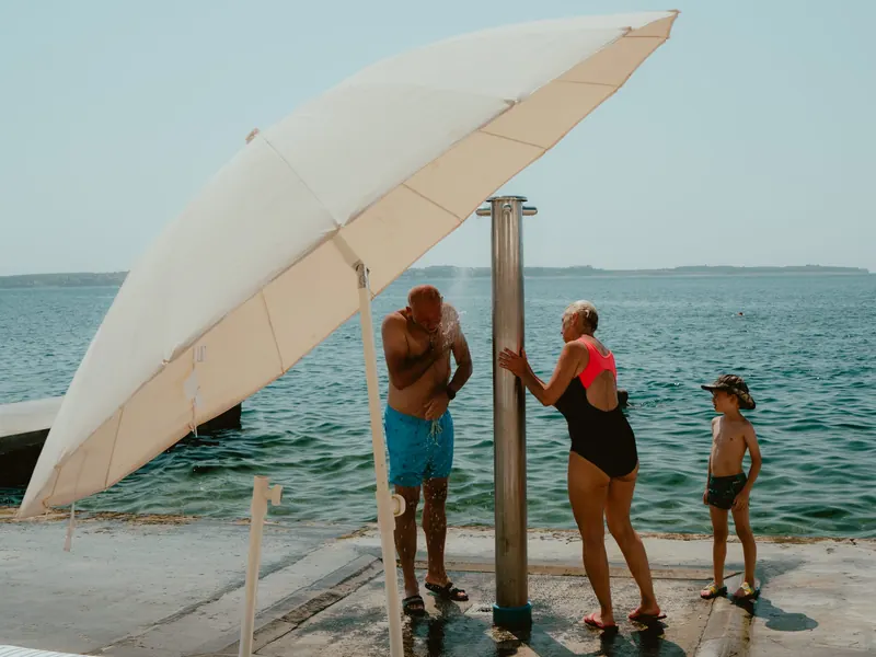 Au bord de la mer, un homme et une femme en maillot se rincent sous une douche de plage, à côté d’un grand parasol blanc ; un petit garçon en casquette attend près d’eux.