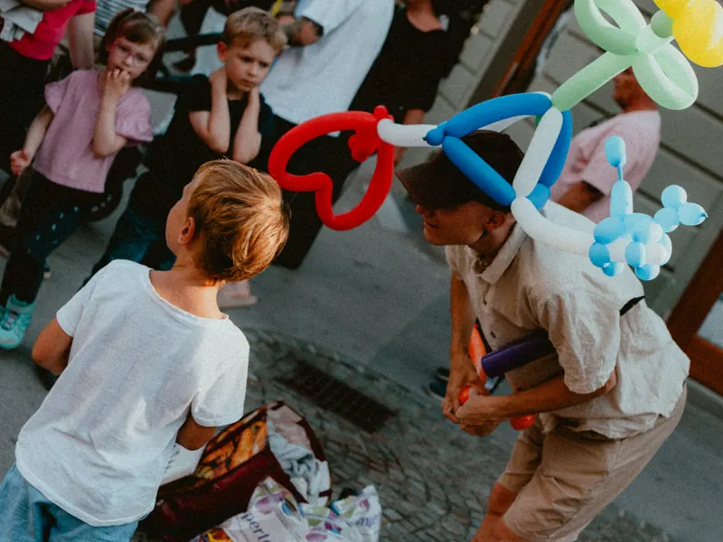 Des enfants regardent un sculpteur de ballons coiffé d’un chapeau en ballons colorés, en forme de cœur, de fleur et d’animal.
