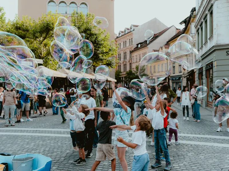Sur une place pavée bordée de façades, des enfants lèvent les bras vers de grandes bulles de savon qui flottent au-dessus des passants.