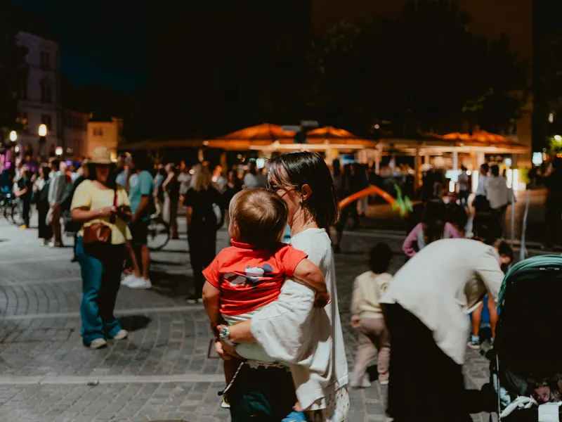 Le soir, sur une place pavée pleine de monde, une femme porte dans ses bras un petit enfant en t-shirt rouge, devant des parasols éclairés.