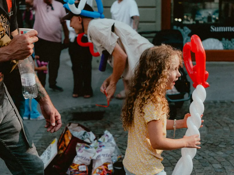 Dans une rue animée, une fillette aux cheveux bouclés tient une longue sculpture en ballons rouge et blanc, à côté du sculpteur penché sur ses sacs.