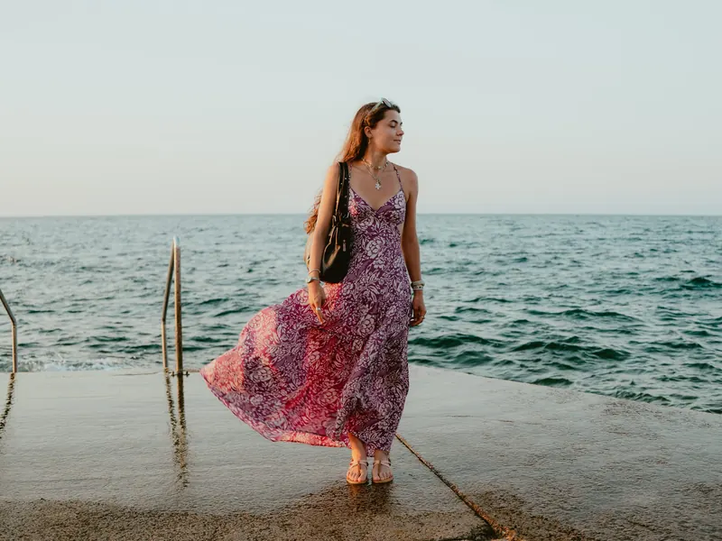 Sur un quai de baignade, une jeune femme en longue robe rose à motifs, que le vent soulève, regarde au loin, près d’une échelle qui descend dans la mer.