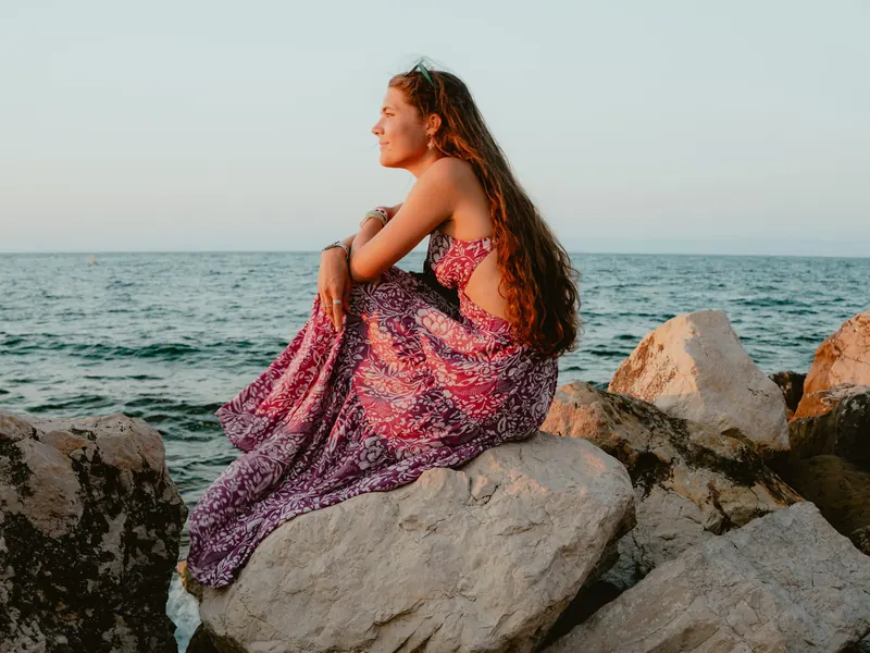 Une jeune femme en robe longue rose et violette, assise sur de gros rochers au bord de la mer, regarde au loin dans la lumière de fin de journée.