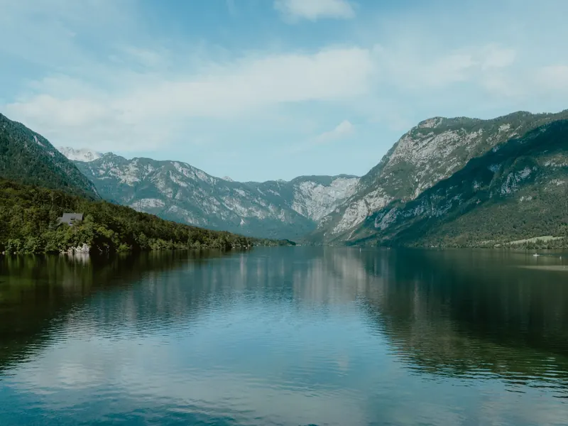 Un lac calme reflète les montagnes boisées qui l’encadrent de part et d’autre, sous un ciel bleu traversé de nuages.