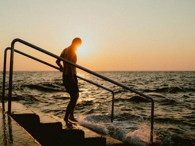 À contre-jour, un homme descend les marches d’un escalier de baignade, une main sur la rampe, face à la mer et au soleil couchant.