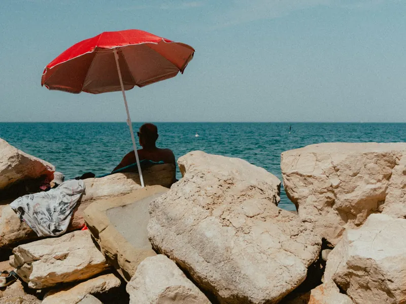 Un parasol rouge planté entre de gros rochers clairs, au bord d’une mer bleue ; derrière les rochers, un homme assis regarde l’eau.
