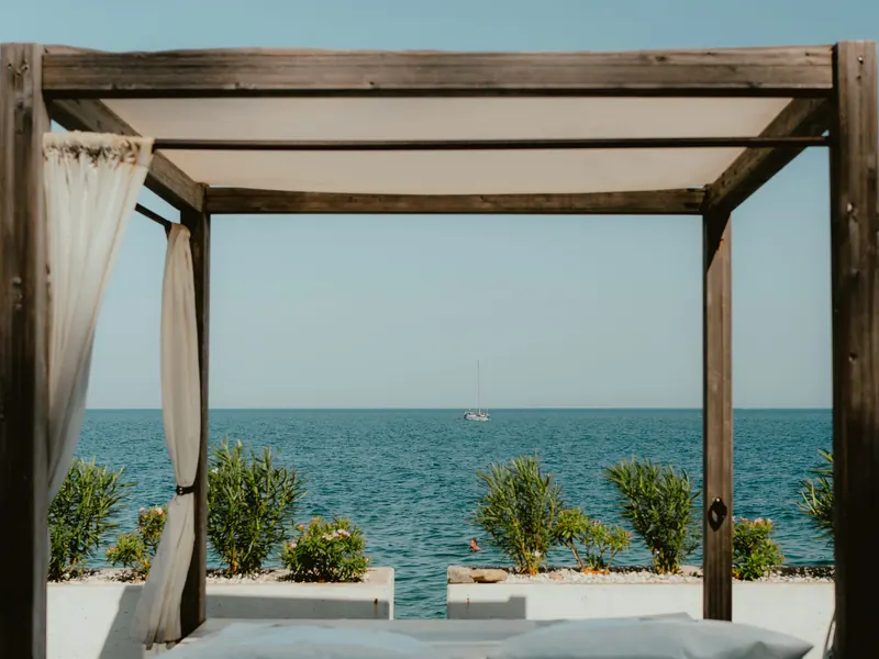 Une pergola en bois aux voilages blancs encadre la mer, un voilier au loin et une rangée de plantes en bacs.
