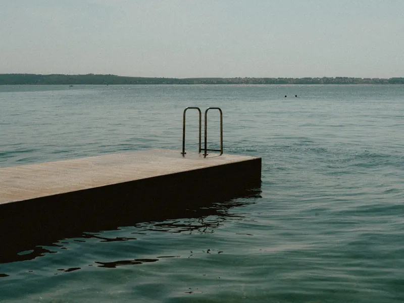 Un ponton avec une échelle de baignade dans une mer calme et laiteuse, sous un ciel voilé ; une côte basse au loin.