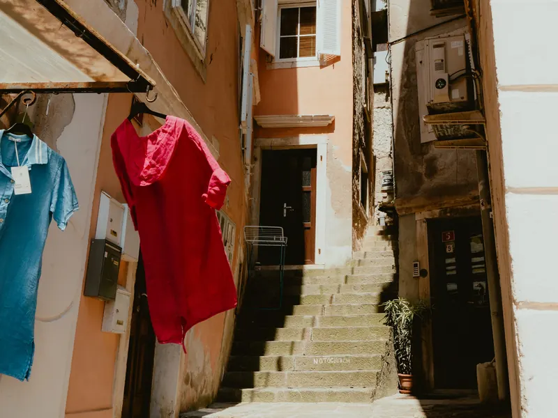 Une ruelle étroite aux façades ocre monte en escalier ; au premier plan, une robe rouge et une chemise bleue sont suspendues devant une boutique.