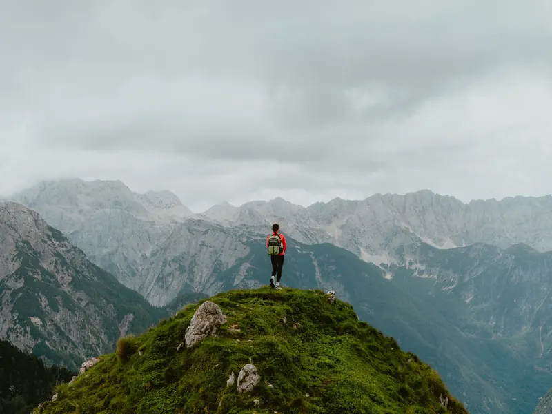 Une personne en veste rouge, debout au sommet d’une crête herbeuse, face à une chaîne de montagnes rocheuses sous un ciel gris.