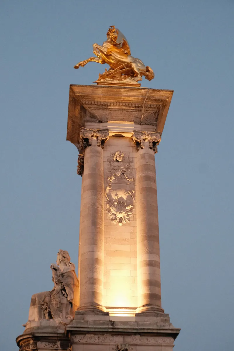 Le haut d’un pilier du pont Alexandre-III, à Paris, éclairé au crépuscule : colonnes de pierre, bas-reliefs et groupe doré avec un cheval ailé sur un ciel bleu.