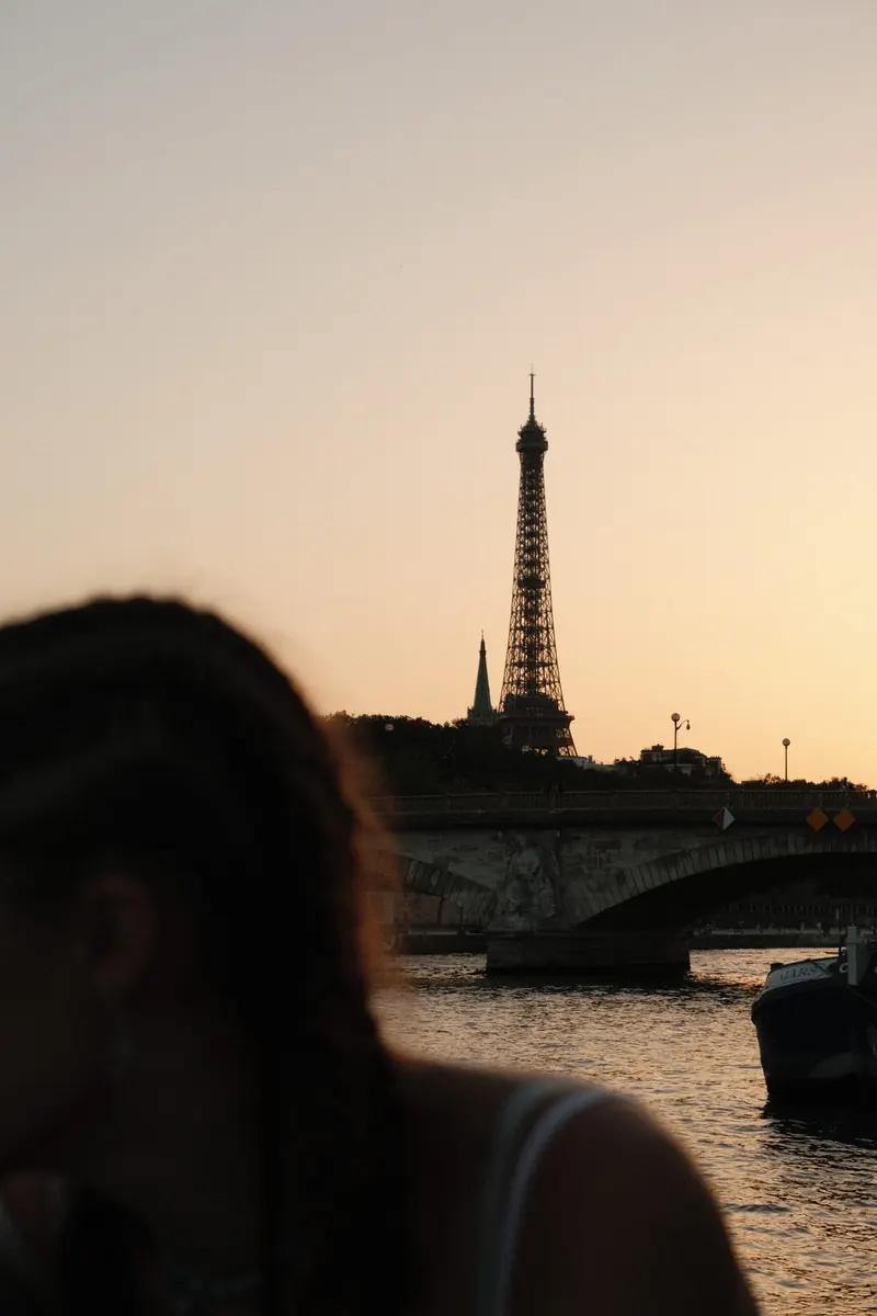 La tour Eiffel se découpe sur un ciel orangé au coucher du soleil, au-dessus d’un pont sur la Seine ; au premier plan, la tête floue d’une personne vue de dos et une péniche amarrée.